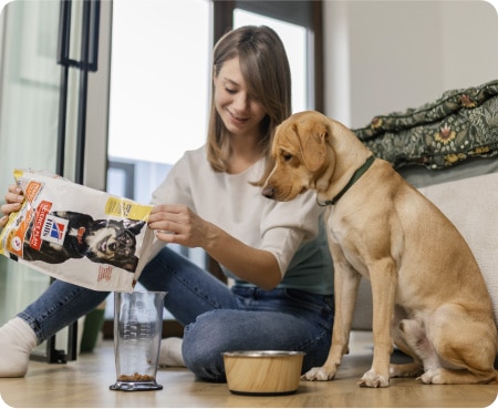 一位女子正在將希爾思寵物營養飼料倒進量杯中，一隻狗在旁邊專注地看著。